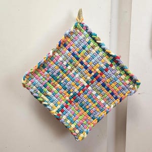 May include: A colorful, woven potholder with a diamond shape. The potholder is made of various colors, including blue, green, yellow, and red. It is hanging on a wall with a gold-colored hook.