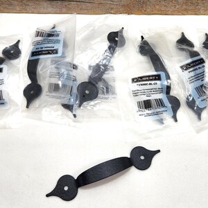 May include: Black wrought iron pull handles with a decorative design. The handles are in clear plastic packaging with product information labels. The handles have a curved shape with decorative ends and are designed for doors or drawers.