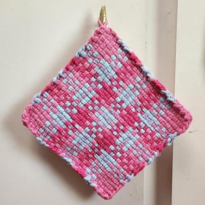 May include: A handmade, square hot pad in shades of pink and light blue, woven in a diagonal pattern. The edges are finished with a pink border. A gold-colored hook is attached to the top corner for hanging.