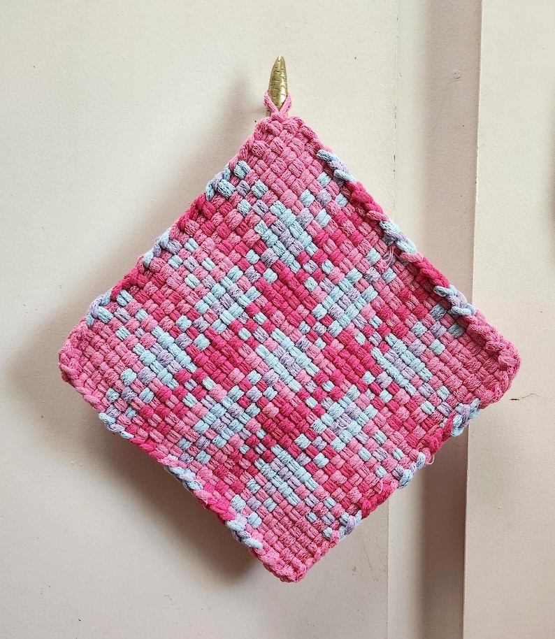 May include: A handmade, square hot pad in shades of pink and light blue, woven in a diagonal pattern. The edges are finished with a pink border. A gold-colored hook is attached to the top corner for hanging.