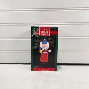 May include: A Hallmark Keepsake Ornament box featuring a North Pole Nutcracker named Frieda, The Animals' Friend. The box displays a nutcracker figure wearing a blue hat, white fur collar, light blue scarf, and a red polka dot apron holding a yellow bird.