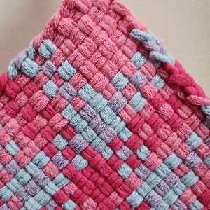 May include: A handmade, woven hot pad in a square shape. The color scheme includes shades of pink, light blue, and purple, arranged in a checkered pattern. A loop is attached at the top for hanging. The texture appears soft and the item is likely made of yarn.