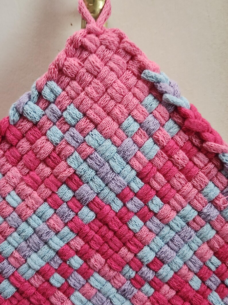 May include: A handmade, woven hot pad in a square shape. The color scheme includes shades of pink, light blue, and purple, arranged in a checkered pattern. A loop is attached at the top for hanging. The texture appears soft and the item is likely made of yarn.