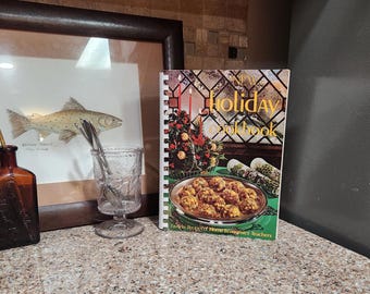 May include: A still-life arrangement featuring a framed fish illustration, a vintage cookbook titled "New Holiday Cookbook," a decorative glass, and a brown bottle with a stopper. The cookbook displays a festive holiday scene.