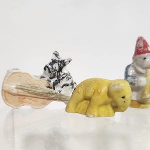 May include: Three small ceramic figurines including a black and white dog playing a tan cello, a yellow elephant, and a white figure wearing a red hat and blue coat while playing a yellow instrument. These miniature items are painted.