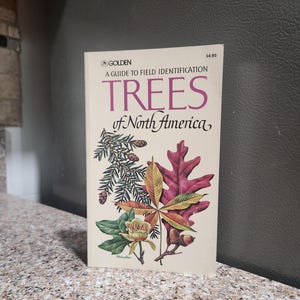 May include: A softcover book titled A Guide to Field Identification Trees of North America by Golden. The off-white cover features detailed botanical illustrations of pine needles with cones, a yellow leaf, a red oak leaf, and a flower.
