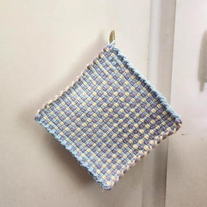 May include: A square, woven potholder in shades of blue, lavender, and yellow. The potholder has a loop for hanging and is made of a tightly woven fabric. The edges are finished with a blue border.