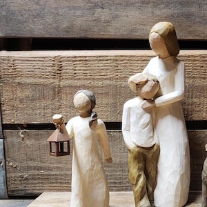 May include: Wooden figurines depicting a family. A woman in a long white dress embraces a child in a white shirt and brown pants. Another figure holds a brown lantern. The figures are set against a wooden background.