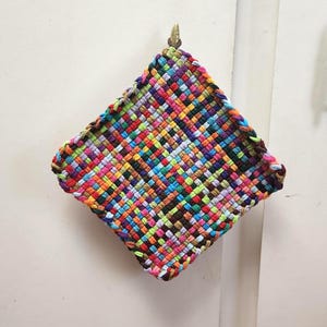 May include: A colorful, woven hot pad with a diamond shape. The pad is made of various colors, including red, orange, yellow, green, blue, and purple. It is hanging on a wall. The edges are a darker brown.
