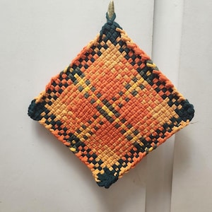 May include: A woven, square hot pad in shades of orange, yellow, and black. The handmade pot holder has a diamond pattern and a dark green border. A small metal loop is attached for hanging.