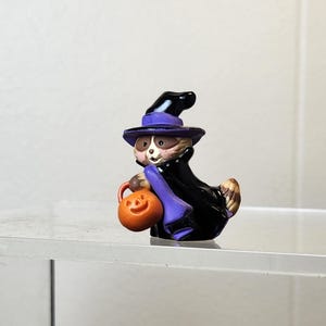 May include: A Halloween-themed figurine of a raccoon dressed as a witch, wearing a black cloak and purple hat. The raccoon holds an orange pumpkin-shaped bag. The figurine is on a clear surface.
