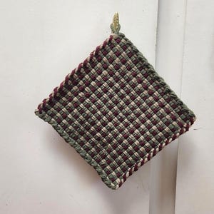 May include: A square, woven potholder in a checkerboard pattern of burgundy, olive green, and cream. The edges are trimmed with a burgundy border. A small, gold-colored hook is attached for hanging.