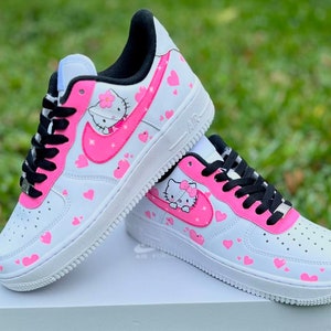May include: White low-top sneakers with a pink Hello Kitty design and pink hearts. The sneakers have black laces and a black swoosh.
