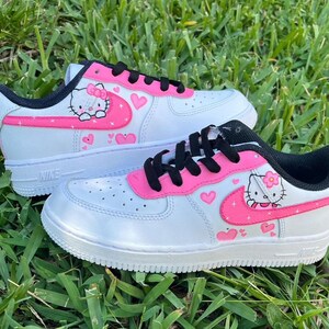 May include: White low-top sneakers with a pink Hello Kitty design. The sneakers have a pink swoosh with hearts and Hello Kitty characters on the side.