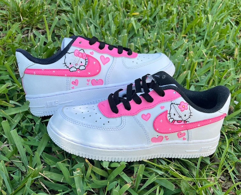 May include: White low-top sneakers with a pink Hello Kitty design. The sneakers have a pink swoosh with hearts and Hello Kitty characters on the side.