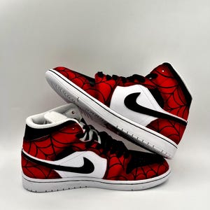 Puede incluir: Dos zapatillas de ca&ntilde;a alta con un dise&ntilde;o de telara&ntilde;a roja y negra. Las zapatillas tienen detalles en blanco, incluyendo las suelas y un panel blanco con un logotipo de Nike negro. Sobre un fondo blanco.