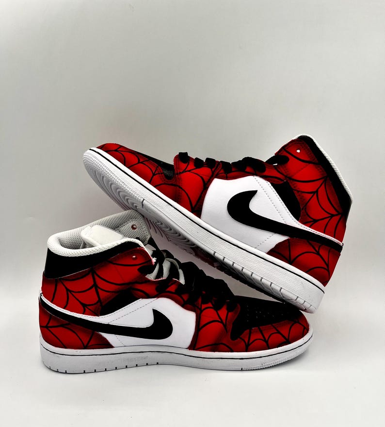 Puede incluir: Dos zapatillas de ca&ntilde;a alta con un dise&ntilde;o de telara&ntilde;a roja y negra. Las zapatillas tienen detalles en blanco, incluyendo las suelas y un panel blanco con un logotipo de Nike negro. Sobre un fondo blanco.