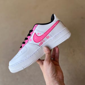 May include: White low-top sneakers with a pink swoosh and pink hearts on the side.