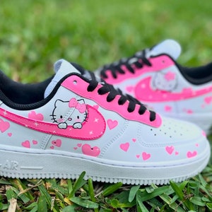 May include: White low-top sneakers with a pink Hello Kitty design and pink hearts. The sneakers have black laces and a black swoosh with a pink outline.