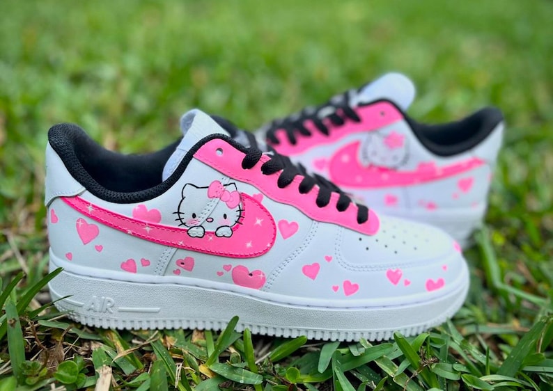 May include: White low-top sneakers with a pink Hello Kitty design and pink hearts. The sneakers have black laces and a black swoosh with a pink outline.