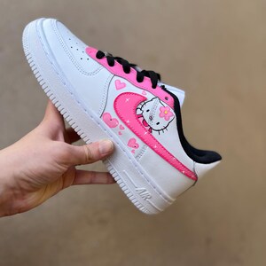 May include: White low-top sneakers with a pink Hello Kitty design. The design features Hello Kitty with a flower, hearts, and stars. The sneakers have a black swoosh and a pink outline.