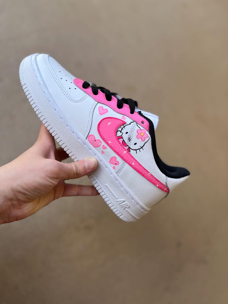May include: White low-top sneakers with a pink Hello Kitty design. The design features Hello Kitty with a flower, hearts, and stars. The sneakers have a black swoosh and a pink outline.