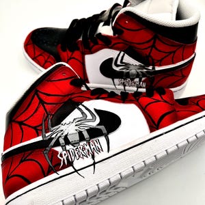 Puede incluir: Par de zapatillas altas con tem&aacute;tica de Spider-Man. Los zapatos presentan una combinaci&oacute;n de colores rojo y negro con un dise&ntilde;o de telara&ntilde;a. El logo de Nike es blanco y la palabra "SPIDERMAN" est&aacute; impresa en el lateral.