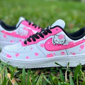 May include: White sneakers with a pink Hello Kitty design and pink hearts. The sneakers have a black swoosh and black laces.