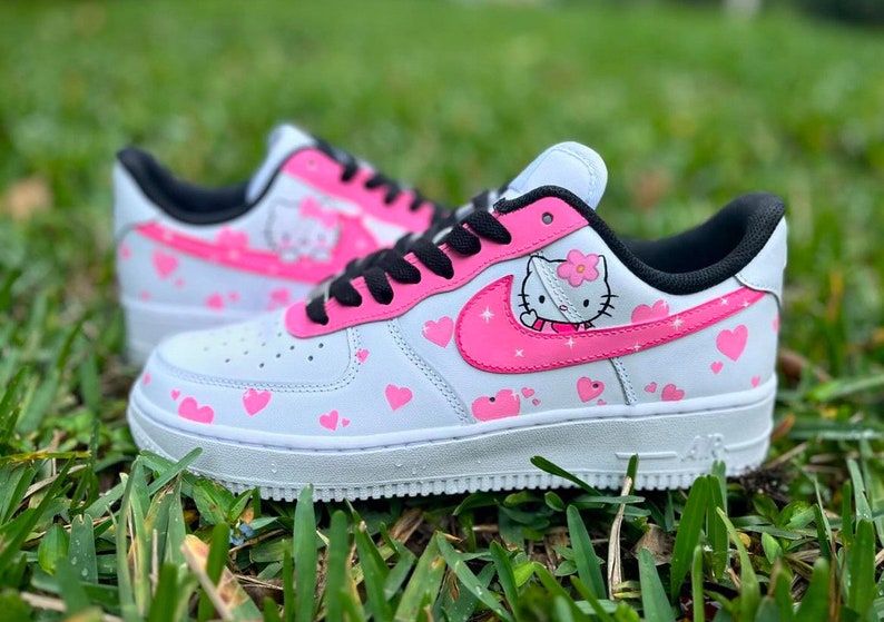May include: White sneakers with a pink Hello Kitty design and pink hearts. The sneakers have a black swoosh and black laces.
