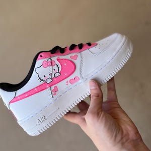 May include: White low-top sneakers with a pink Hello Kitty design on the side. The design features Hello Kitty with a pink bow and a pink heart. The sneakers have a black swoosh and a white sole.