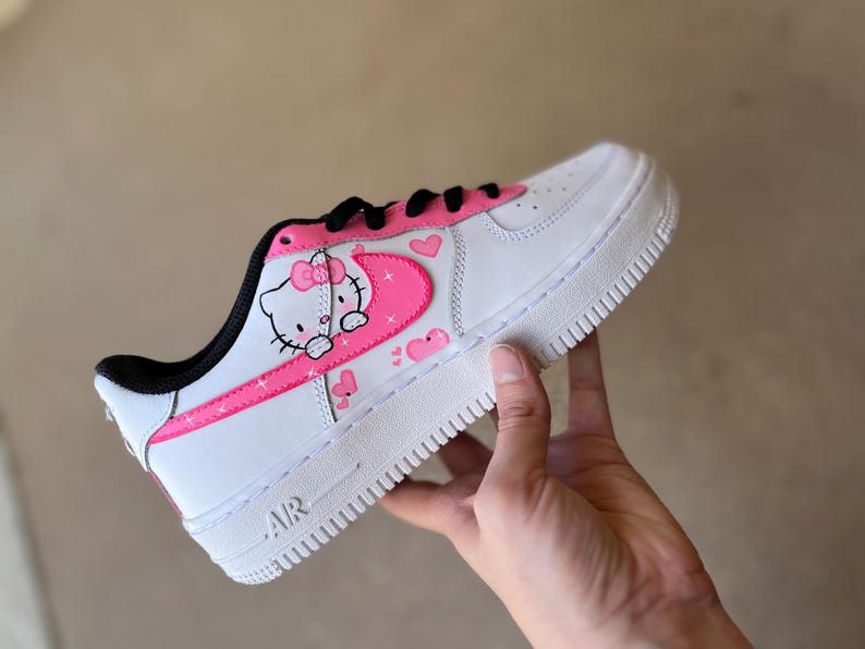 May include: White low-top sneakers with a pink Hello Kitty design on the side. The design features Hello Kitty with a pink bow and a pink heart. The sneakers have a black swoosh and a white sole.