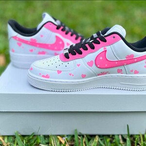 May include: White low-top sneakers with a pink heart pattern and a pink swoosh logo. The sneakers are sitting on a white box.
