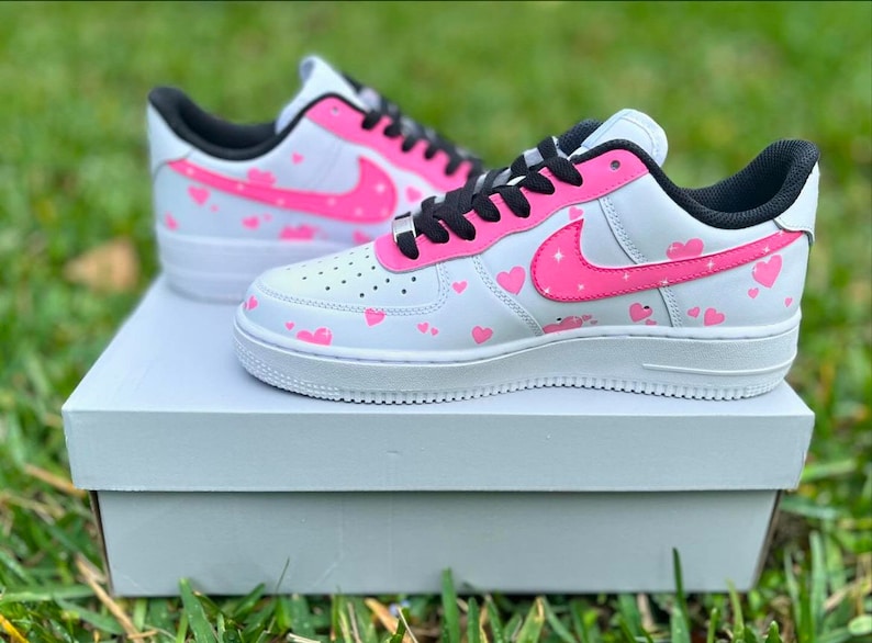 May include: White low-top sneakers with a pink heart pattern and a pink swoosh logo. The sneakers are sitting on a white box.