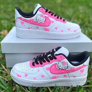 May include: White low-top sneakers with pink hearts and a Hello Kitty design on the side. The sneakers have black laces and a black swoosh.