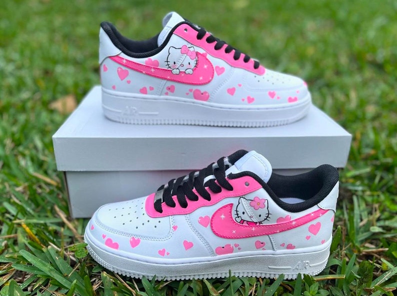 May include: White low-top sneakers with pink hearts and a Hello Kitty design on the side. The sneakers have black laces and a black swoosh.