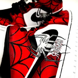 Puede incluir: Par de zapatillas altas con un dise&ntilde;o de telara&ntilde;a rojo y negro. Las zapatillas presentan un logotipo de Nike blanco y un gr&aacute;fico de ara&ntilde;a con la palabra "SPIDERMAN". Los zapatos tienen suelas blancas y cordones negros.