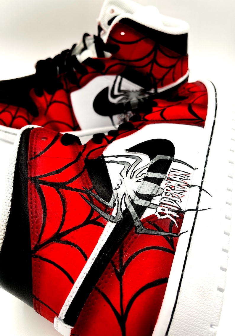 Puede incluir: Par de zapatillas altas con un dise&ntilde;o de telara&ntilde;a rojo y negro. Las zapatillas presentan un logotipo de Nike blanco y un gr&aacute;fico de ara&ntilde;a con la palabra "SPIDERMAN". Los zapatos tienen suelas blancas y cordones negros.