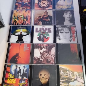 May include: A collection of 12 compact discs (CDs) in jewel cases. The CDs are arranged in three rows of four. The CDs feature a variety of artists and album titles, including "U2", "The Tea Party", "Edwin", "Born Again", "Live", "In Da House", "Mr. Bungle", "Hold", and "Our Time" by Bryan Adams.