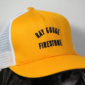 May include: A yellow and white trucker hat with the text "Ray Gouge Firestone" embroidered in black.