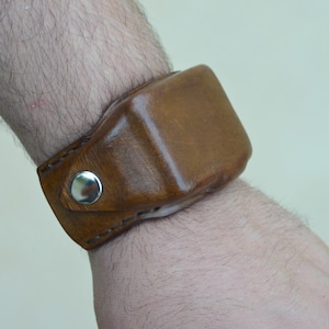 Könnte beinhalten: Ein braunes Lederarmband mit rechteckigem Design. Das Armband wird mit einem silbernen Druckknopfverschluss befestigt. Das Leder hat eine leicht glänzende Oberfläche und weist natürliche Farbvariationen auf.