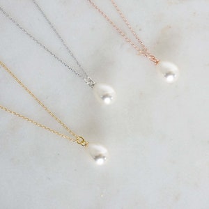 May include: Four delicate necklaces with teardrop-shaped white pearls. The chains are gold, silver, and rose gold. The necklaces are displayed on a white marble surface, creating a minimalist and elegant aesthetic.