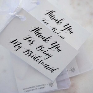 May include: Two white gift tags with black calligraphy that reads "Thank You for Being My Bridesmaid".