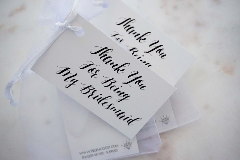 May include: Two white gift tags with black calligraphy that reads "Thank You for Being My Bridesmaid".