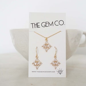 Puede incluir: Un conjunto de collar y pendientes de oro con un colgante en forma de diamante y pendientes a juego. El colgante y los pendientes están adornados con piedras de circonita cúbica transparentes. Las joyas se presentan en una tarjeta blanca con el texto "THE GEM CO." y "www.thegemshop.com".