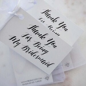 May include: White gift tags with black calligraphy reading "Thank You For Being My Bridesmaid." The tags are tied together with a white ribbon. The tags are on a white surface.