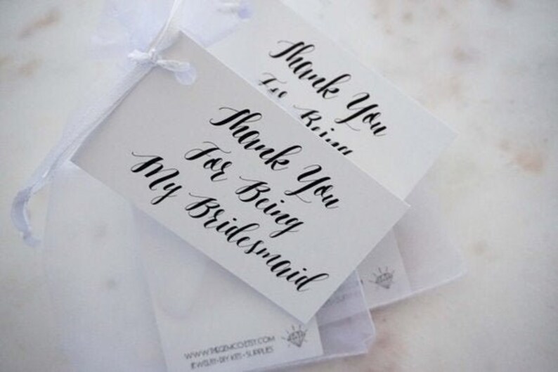May include: White gift tags with black calligraphy reading "Thank You For Being My Bridesmaid." The tags are tied together with a white ribbon. The tags are on a white surface.