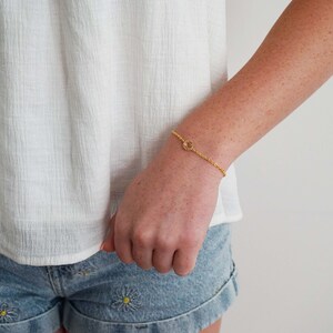 May include: A gold chain bracelet with a small, circular charm. The charm has a textured, sun-like design.