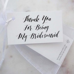 May include: A white gift tag with the text "Thank You For Being My Bridesmaid" in elegant black calligraphy. The tag is tied with a white ribbon and sits atop a white card and a sheer white bag.