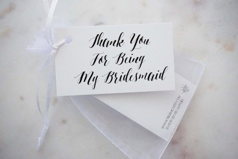 May include: A white gift tag with the text "Thank You For Being My Bridesmaid" in elegant black calligraphy. The tag is tied with a white ribbon and sits atop a white card and a sheer white bag.