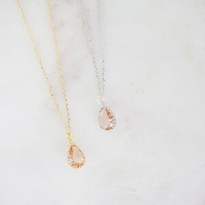 May include: Two delicate necklaces with teardrop-shaped pendants. One necklace has a gold chain and pendant, while the other features a silver chain and pendant. The pendants are a light peach color, with a textured appearance.